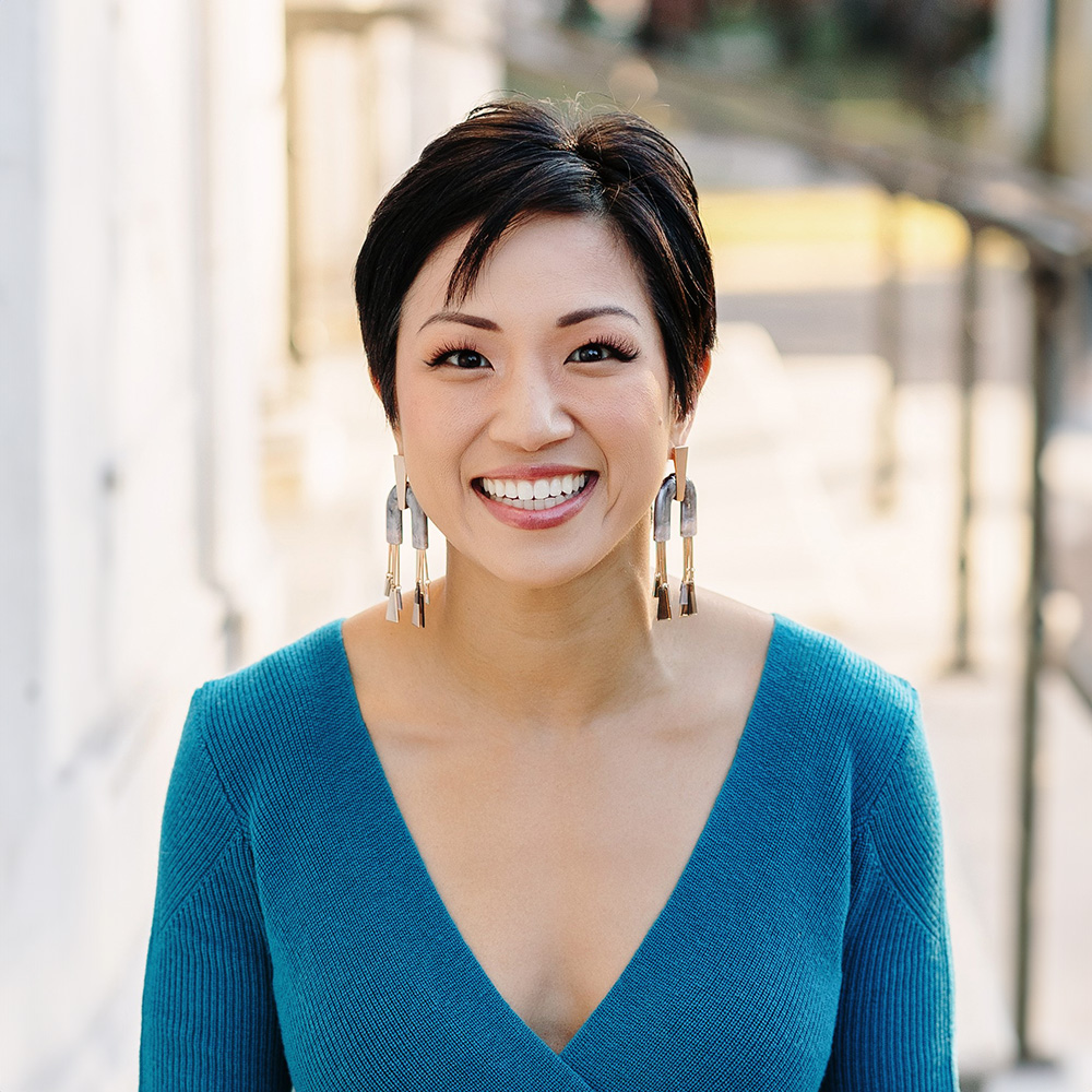 The image shows a person with short hair smiling at the camera, wearing a blue top, standing outdoors on a sunny day.