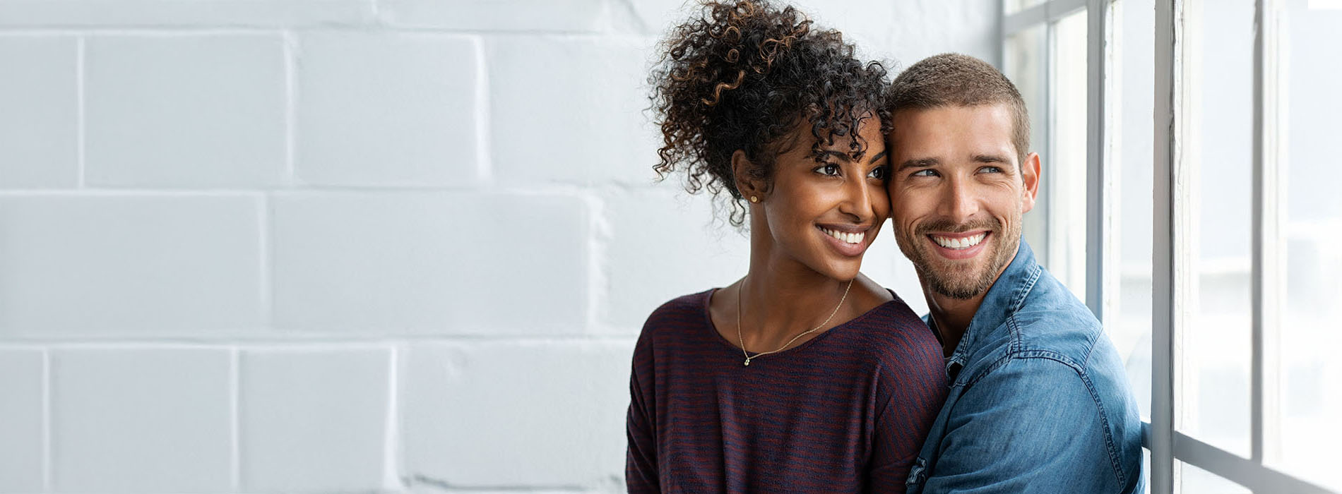 A man and woman, possibly a couple, are standing close together with smiles on their faces, looking at each other, against a white brick wall background.