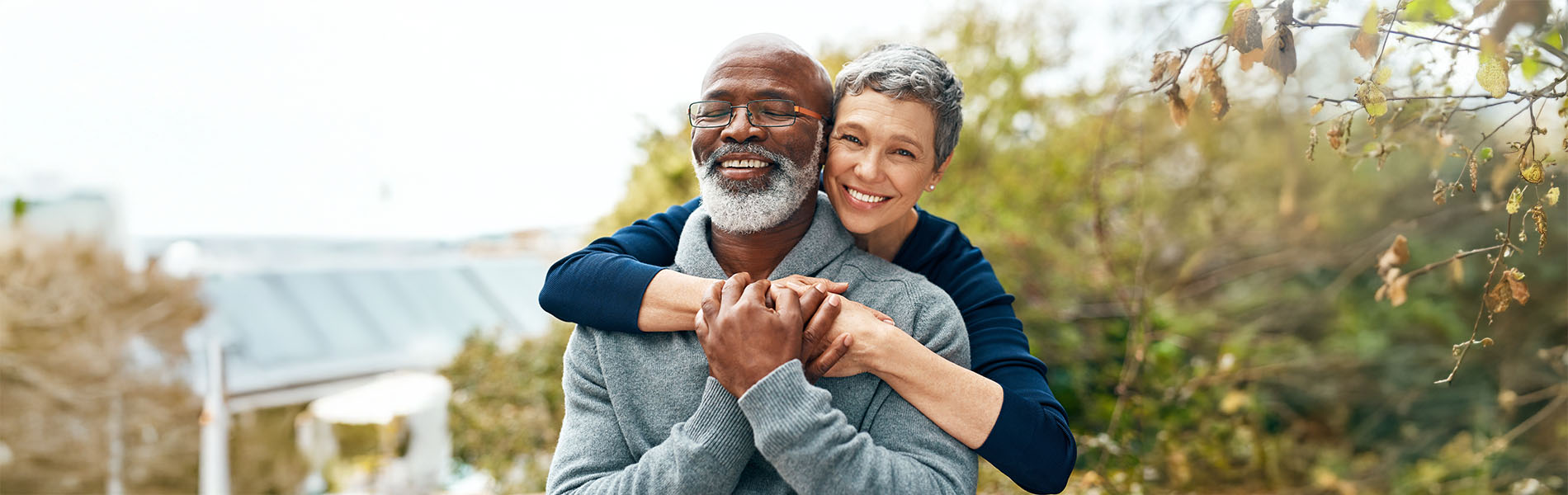 An elderly man and woman are embracing each other outdoors, with trees and foliage in the background.