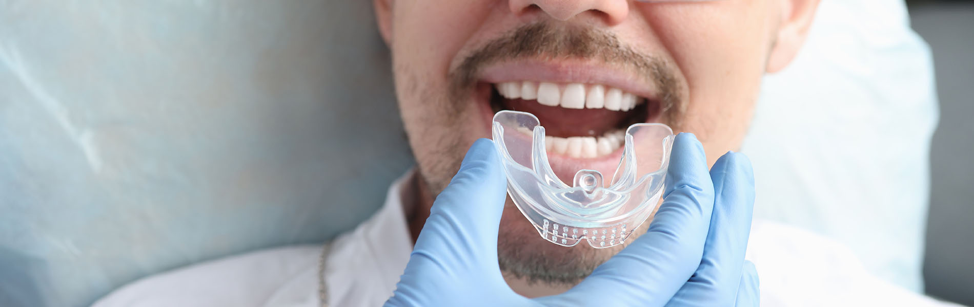 The image depicts a man wearing eyeglasses and a surgical mask, holding a transparent dental impression device up to his mouth with one hand while sitting in a chair, likely in a dental office setting.