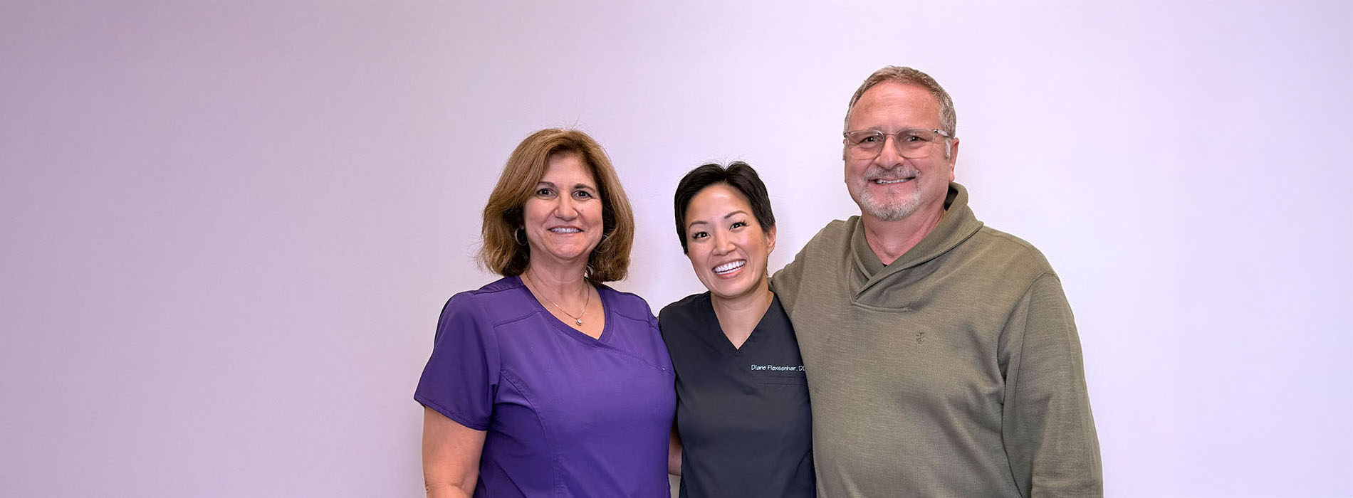 The image shows three individuals posing together with a backdrop  an adult woman, an adult man, and a younger person who appears to be a girl. They are standing side by side, smiling at the camera.