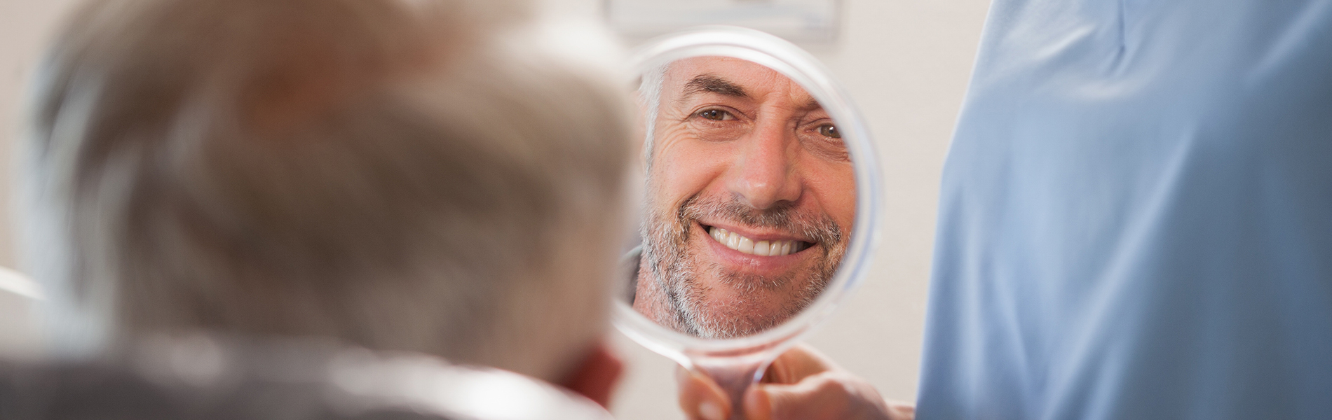 The image shows a man with a beard sitting in front of a mirror, being attended to by a person whose face is obscured by a blue surgical drape.