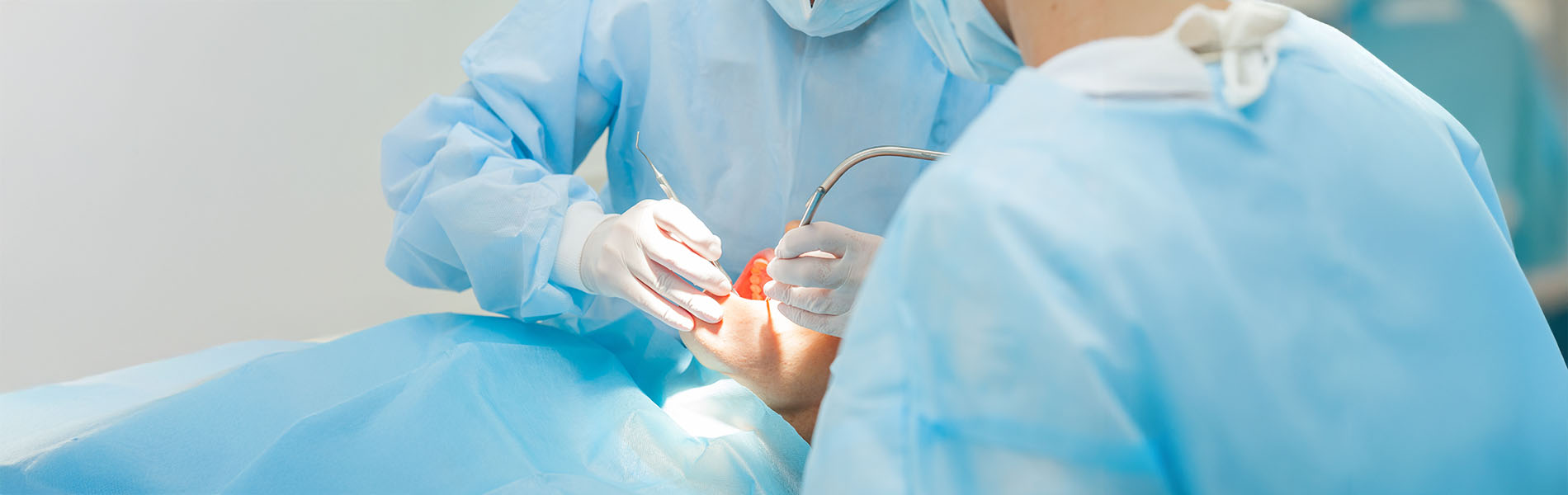 The image shows two individuals wearing surgical gowns and face masks  one person appears to be performing a medical procedure on another, with a focus on the hands and instruments involved.