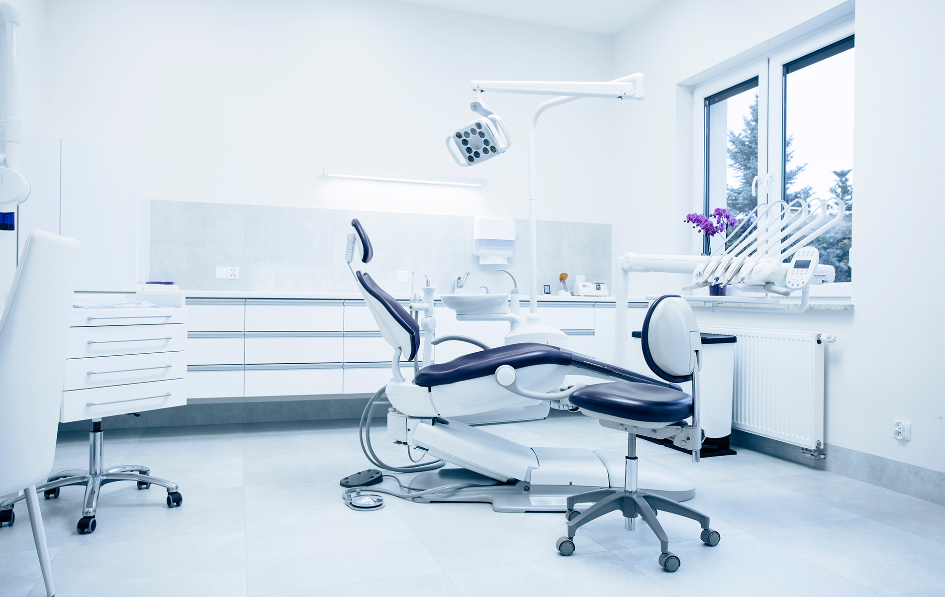 The image displays a modern dental office interior with various equipment and furniture typical of such a setting, including chairs, a workstation, and a large window allowing natural light into the room.