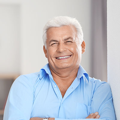 The image features a man with gray hair, wearing a blue shirt, leaning against a wall with his arms crossed, smiling at the camera.