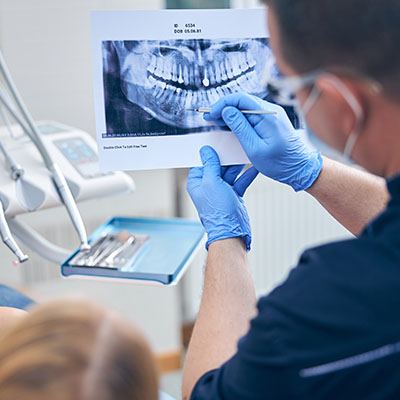 The image depicts a dental professional examining a patient s teeth using a magnifying glass and a dental mirror, with an X-ray displayed on a screen behind them.