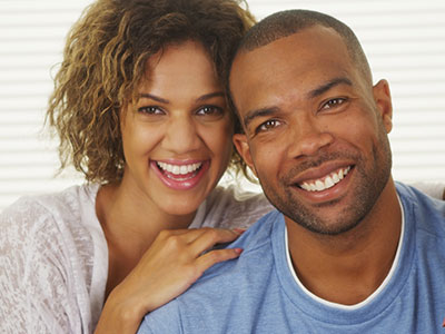 The image shows a man and a woman posing together with smiles, the man wearing a short beard and the woman with curly hair.